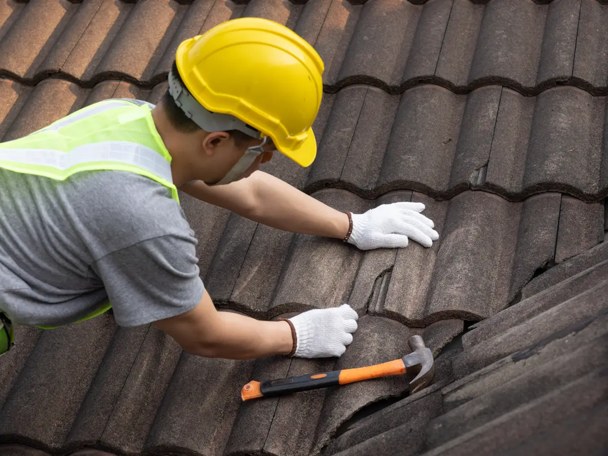 Roofer in a hard hat repairing a tile roof with a hammer