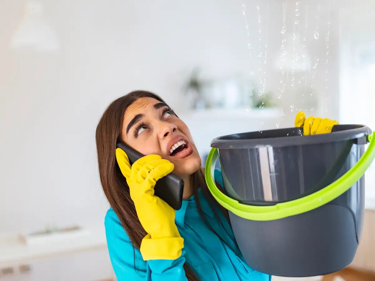 Homeowner on the phone catching water dripping through the ceiling in a bucket